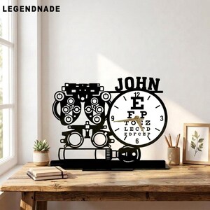 May include: A black metal desk clock featuring a silhouette of an optometry phoropter and a circular clock face designed like a Snellen eye chart. The name JOHN is displayed in black lettering above the clock face, which has gold-colored hands.