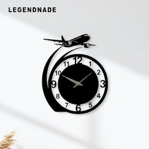 May include: Black wall clock featuring a jet airplane design. The clock face is black with white numerals and silver hands. The airplane is black and appears to be ascending. The word "LEGENDNADE" is printed at the top left.