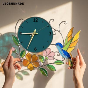 Könnte beinhalten: Eine runde Wanduhr mit dunkelblau-grünem Zifferblatt, goldenen Zeigern und den schwarzen Ziffern 3, 6, 9 und 12. Der Rahmen zeigt Buntglas-Elemente wie einen blauen Kolibri, gelbe und orangefarbene Blumen, rosa Blüten und grüne Blätter.