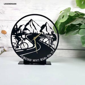 May include: Black circular clock featuring a mountain scene with cyclists on a road. The clock has gold-colored hands and a rectangular base. The text "CRUSH MS!! Ride On !!" is displayed on the clock.