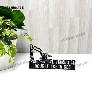 以下が含まれることがあります： 「JON SCHNEIDER DOUBLE J SERVICES」の文字が入った、黒い金属製のショベルカーのサイン。ショベルカーのシルエットです。背景には鉢植えの植物があります。
