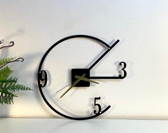 Può includere: Orologio da parete in metallo nero dal design minimalista. L'orologio presenta un telaio circolare con i numeri 3, 5 e 9. Le lancette sono dorate e il quadrante è un quadrato nero. Il marchio "LEGENDNADE" è in alto a sinistra.
