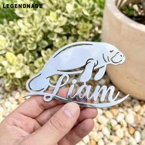 May include: A silver-colored metal manatee-shaped nameplate with the name "Liam" cut out in a cursive font. The nameplate is held in a hand, with the background featuring greenery and a pot.