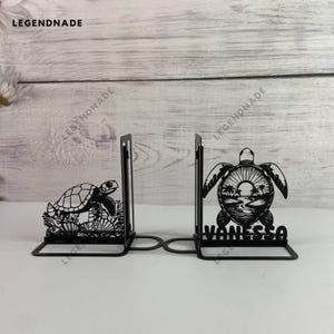 May include: Black metal bookends featuring turtle designs. One bookend depicts a turtle on coral, the other a turtle with a sunset scene and the name "VANESSA". The brand name "LEGENDNADE" is visible.