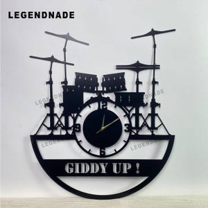 Op de afbeelding: Zwarte metalen wandklok in de vorm van een drumstel, met bekkens, drums en een wijzerplaat. De klok heeft gouden wijzers en de tekst "GIDDY UP!" staat onderaan. De merknaam "LEGENDNADE" staat bovenaan.