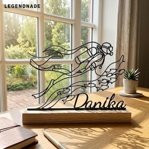 May include: A black metal wire sculpture of a scuba diver swimming with a school of fish, mounted on a rectangular wooden base. The name Danika is cut out in cursive script at the bottom of the metal design. The piece is displayed on a wooden surface.