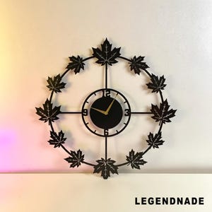 Könnte beinhalten: Schwarze Metall-Wanduhr mit rundem Zifferblatt und goldenen Zeigern. Die Uhr ist von einem Ring aus Ahornblatt-Ausschnitten umgeben. Der Text "LEGENDNADE" befindet sich unten.