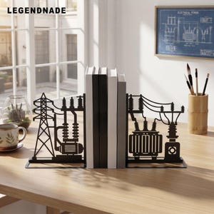 May include: A pair of black metal bookends featuring intricate laser-cut designs of electrical infrastructure, including a transmission tower, a transformer, and power lines, supporting a row of four books on a wooden surface.