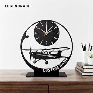 May include: Black metal desk clock with an airplane silhouette and a circular clock face. The clock has gold hands and the text "CUSTOM NAME" is displayed below the airplane. The brand name "LEGENDNADE" is at the top left.