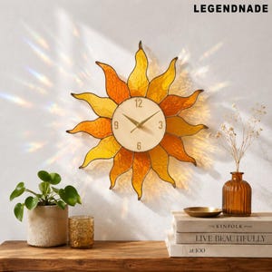 May include: A decorative wall clock shaped like a sun with yellow and orange stained glass rays. The clock face is cream-colored with black numerals and gold hands. The brand name "LEGENDNADE" is printed in the upper right corner.