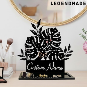 May include: Black jewelry display stand with a monstera leaf design. The stand features a tray for rings and a section for necklaces and earrings. The text "Custom Name" is displayed on the front. The brand name "LEGENDNADE" is at the top.