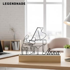 Puede incluir: Un dibujo lineal en blanco y negro de una mujer tocando un piano de cola, montado sobre una base de madera. El nombre "Anna" está escrito en cursiva. La palabra "LEGENDNADE" está en la esquina superior izquierda.
