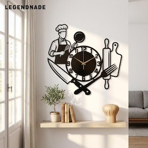 May include: A black metal wall clock featuring a chef silhouette, a large knife, a fork, a rolling pin, and a cutting board. The clock face has gold-coloured hands and black numbers. The text LEGENDNADE is visible in the upper left corner.