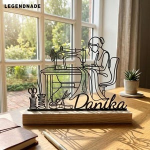 Peut inclure: Une sculpture en fil de métal noir représentant une personne assise devant une machine à coudre, accompagnée de ciseaux, de bobines de fil et d'un ruban à mesurer. Le nom Danika est écrit en lettres cursives sur un socle en bois rectangulaire.