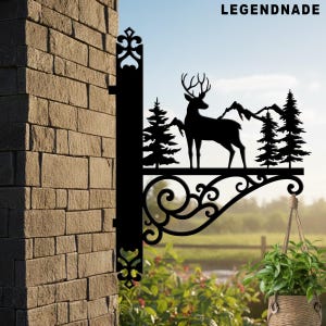 May include: Black metal hanging sign featuring a deer silhouette with antlers, trees, and mountains. The sign is mounted on a brick wall and includes the text "LEGENDNADE". A decorative plant in a woven basket hangs below.