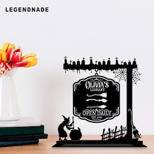 May include: Black metal Halloween-themed sign with the text "Olivia's Library" and "Open Daily Est. 2025". The sign features a witch, cauldron, tombstones, cobwebs, and a broom. The sign is on a wooden surface.