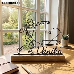 Pode incluir: Uma escultura de fio de metal preto representando uma pessoa a lançar o peso, montada numa base retangular de madeira. A escultura inclui o nome Danika em escrita cursiva e um elemento circular. A palavra LEGENDNADE é visível no canto superior esquerdo.