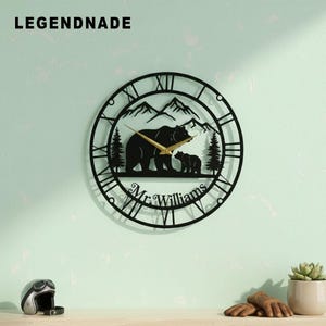 May include: Black metal wall clock with a bear family silhouette, mountains, and Roman numerals. The clock hands are gold. The text "Mr. Williams" is at the bottom. The brand name "LEGENDNADE" is at the top left.
