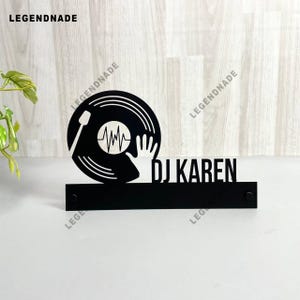 Puede incluir: Letrero de DJ de metal negro con un diseño de disco de vinilo, una silueta de mano y un gráfico de onda de sonido. El nombre "DJ KAREN" se muestra en negrita y mayúsculas. El letrero está montado sobre una base rectangular.