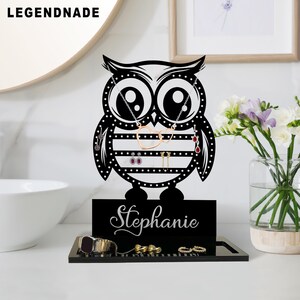 May include: Black owl-shaped jewellery organiser with the name "Stephanie" on the base. The organiser holds necklaces, earrings, and rings. The owl has a decorative design with white dots and horizontal stripes. The brand name "LEGENDNADE" is in the top left corner.