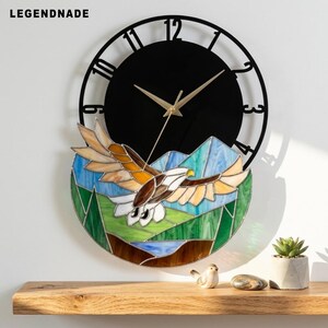 Könnte beinhalten: Eine runde Wanduhr mit dem Text LEGENDNADE in der oberen linken Ecke. Die Uhr hat ein schwarzes Zifferblatt mit goldenen Zeigern und einen unteren Bereich aus Buntglas, der einen fliegenden Adler über grünen Bergen und einem blauen Fluss zeigt.