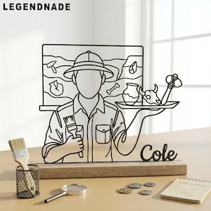 Pode incluir: Escultura de arame preto de um arqueólogo segurando um pincel e uma bandeja com artefatos. A escultura está em uma base de madeira com o nome "Cole". Um pincel, uma lupa, moedas e um bloco de notas também estão presentes.