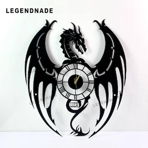 May include: Black dragon-shaped wall clock with a circular clock face featuring Roman numerals. The clock has a dragon head and wings design, with the text "LIZA" at the bottom. The brand name "LEGENDNADE" is at the top.