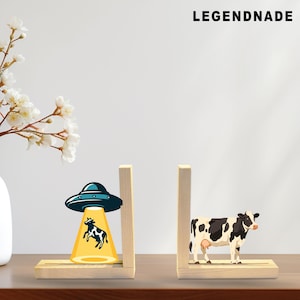 May include: Wooden bookends featuring a cartoon cow and a UFO beaming up a cow. The bookends are made of light-colored wood and have a rectangular base. The text "LEGENDNADE" is at the top.