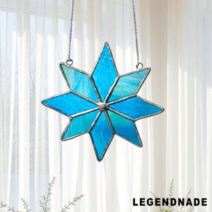 May include: A stained glass star in shades of blue and teal, hanging from a silver chain. The star has eight points and is made of individual glass pieces. The word "LEGENDNADE" is printed in black at the bottom.