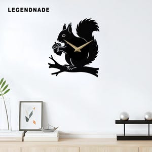 May include: Black squirrel wall clock with gold-colored hands, holding an acorn while perched on a branch. The clock has the word "LEGENDNADE" in white text at the top left. A decorative piece for a living room or bedroom.