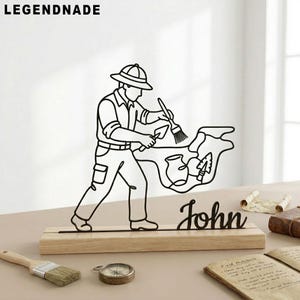 Peut inclure: Sculpture en métal noir représentant un archéologue avec un pinceau et une truelle, montée sur une base en bois. Le nom "John" est écrit en cursive en dessous. Le mot "LEGENDNADE" est en haut.