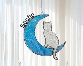 May include: A stained glass cat silhouette sits on a blue crescent moon, suspended by a chain. The word "Sache" is above the moon. The design is set against a window. The word "LEGENDNADE" is at the bottom.
