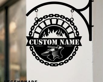 May include: Black metal sign with a blacksmith theme. The sign features a circular design with a chain border, a banner that reads "CUSTOM NAME", and an anvil with a hammer. The sign is suspended from a decorative bracket.