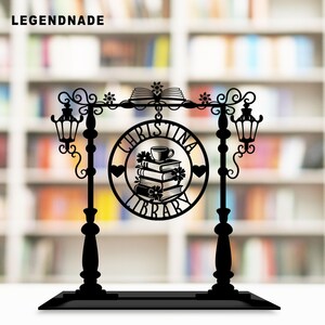 May include: Black metal sign with the text "CHRISTINA LIBRARY" in a circular design, featuring stacked books, a teacup, and floral accents. The sign is supported by two lamp posts and a base, with the word "LEGENDNADE" at the top.