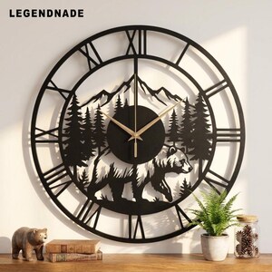 Könnte beinhalten: Eine große, runde, schwarze Wanduhr mit dem Schriftzug LEGENDNADE oben. Das Zifferblatt zeigt ein ausgeschnittenes Design eines Bären, der durch einen Wald vor einer Bergkulisse läuft. Sie hat römische Ziffern und dünne goldene Uhrzeiger.