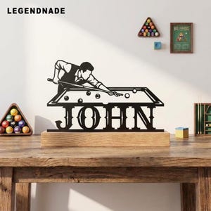 Peut inclure: Une enseigne en métal noir découpée représentant la silhouette d'une personne jouant au billard au-dessus du nom JOHN en grandes lettres capitales, montée sur une base rectangulaire en bois clair. Le design inclut un billard avec des boules.