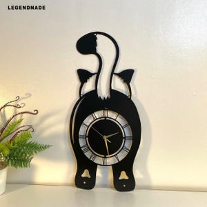 Puede incluir: Reloj de pared negro con forma de gato con cola y orejas blancas. La esfera del reloj es negra con manecillas doradas y números romanos. El reloj es de metal y tiene el texto "LEGENDNADE" en la parte superior izquierda.