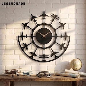 Könnte beinhalten: Eine schwarze, kreisförmige Wanduhr mit einem zentralen Zifferblatt mit dünnen Zeigern und einem Rahmen, der mit Silhouetten verschiedener Flugzeuge in einem radialen Muster verziert ist. Der Text LEGENDNADE steht oben links im Bild.