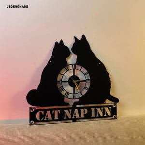 Puede incluir: Reloj de pared de metal negro con dos siluetas de gatos, una esfera con n&uacute;meros romanos y el texto "CAT NAP INN". El reloj tiene manecillas doradas y est&aacute; dise&ntilde;ado para montaje en pared.