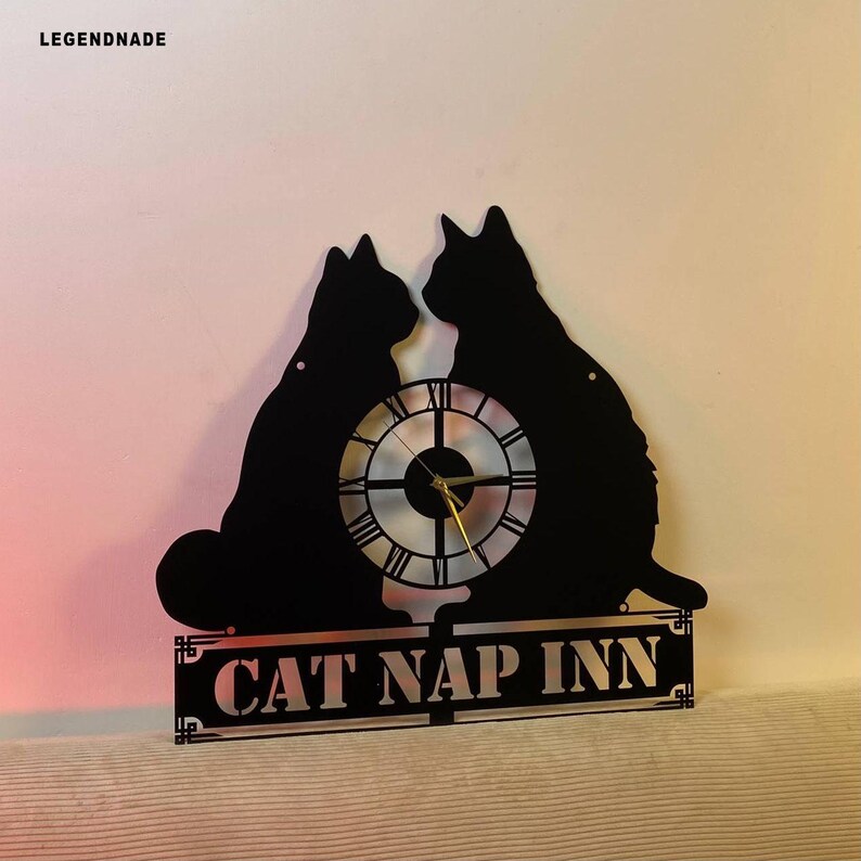 Puede incluir: Reloj de pared de metal negro con dos siluetas de gatos, una esfera con n&uacute;meros romanos y el texto "CAT NAP INN". El reloj tiene manecillas doradas y est&aacute; dise&ntilde;ado para montaje en pared.
