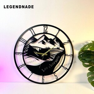 May include: Black metal wall clock with a mountain landscape design. The clock face features Roman numerals and hands, with the text "Les charmes" and the brand name "LEGENDNADE". The clock is round and has a decorative, open design.