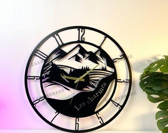May include: Black metal wall clock with a mountain landscape design. The clock face features Roman numerals and hands, with the text "Les charmes" and the brand name "LEGENDNADE". The clock is round and has a decorative, open design.