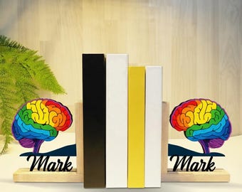 Puede incluir: Soportes para libros de madera con un diseño de cerebro arcoíris y el nombre "Mark" en escritura negra. Los sujetalibros sostienen una fila de libros en negro, blanco y amarillo. La palabra "LEGENDNADE" está impresa en la parte inferior.