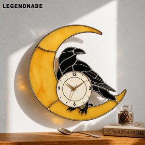 Peut inclure: Une horloge murale décorative représentant un corbeau noir perché sur un croissant de lune jaune. Le cadran de l'horloge est de couleur crème avec des chiffres noirs et des aiguilles dorées. La marque "LEGENDNADE" est visible.