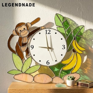 May include: A stained glass wall clock featuring a brown monkey, green leaves, and yellow bananas surrounding a white circular clock face with black numbers 3, 6, 9, and 12. The text LEGENDNADE is printed in the upper left corner of the image.