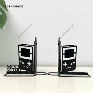 Puede incluir: Dos sujetalibros de metal negro con forma de consola de videojuegos portátil retro. Los sujetalibros tienen una pantalla, botones y el texto "MOD RETRO".