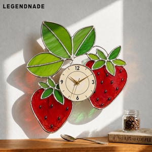 Op de afbeelding: Een decoratieve wandklok met een aardbeienontwerp. De wijzerplaat is crèmekleurig en bevindt zich tussen twee rode aardbeien met groene bladeren. De wijzers van de klok zijn goudkleurig. De merknaam "LEGENDNADE" staat bovenaan.