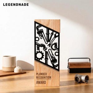 May include: A rectangular wooden award plaque featuring a black metal cutout insert with plumbing tool silhouettes including wrenches, gears, and pipes. The text PLUMBER RECOGNITION AWARD is printed in black on the lower wooden section of the plaque.