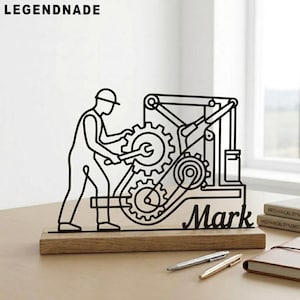 May include: Black metal sculpture of a mechanic working on gears, mounted on a wooden base. The name "Mark" is cut out below the gears. The word "LEGENDNADE" is at the top. Pens and a notebook are on the table.
