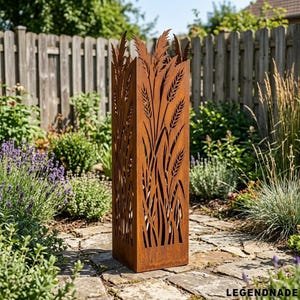 Può includere: Una scultura da giardino alta e rettangolare in metallo ossidato con disegni di spighe di grano ritagliate. La scultura è di colore marrone ruggine e si trova su un sentiero di pietra in un giardino, circondata da piante verdi e una staccionata di legno.